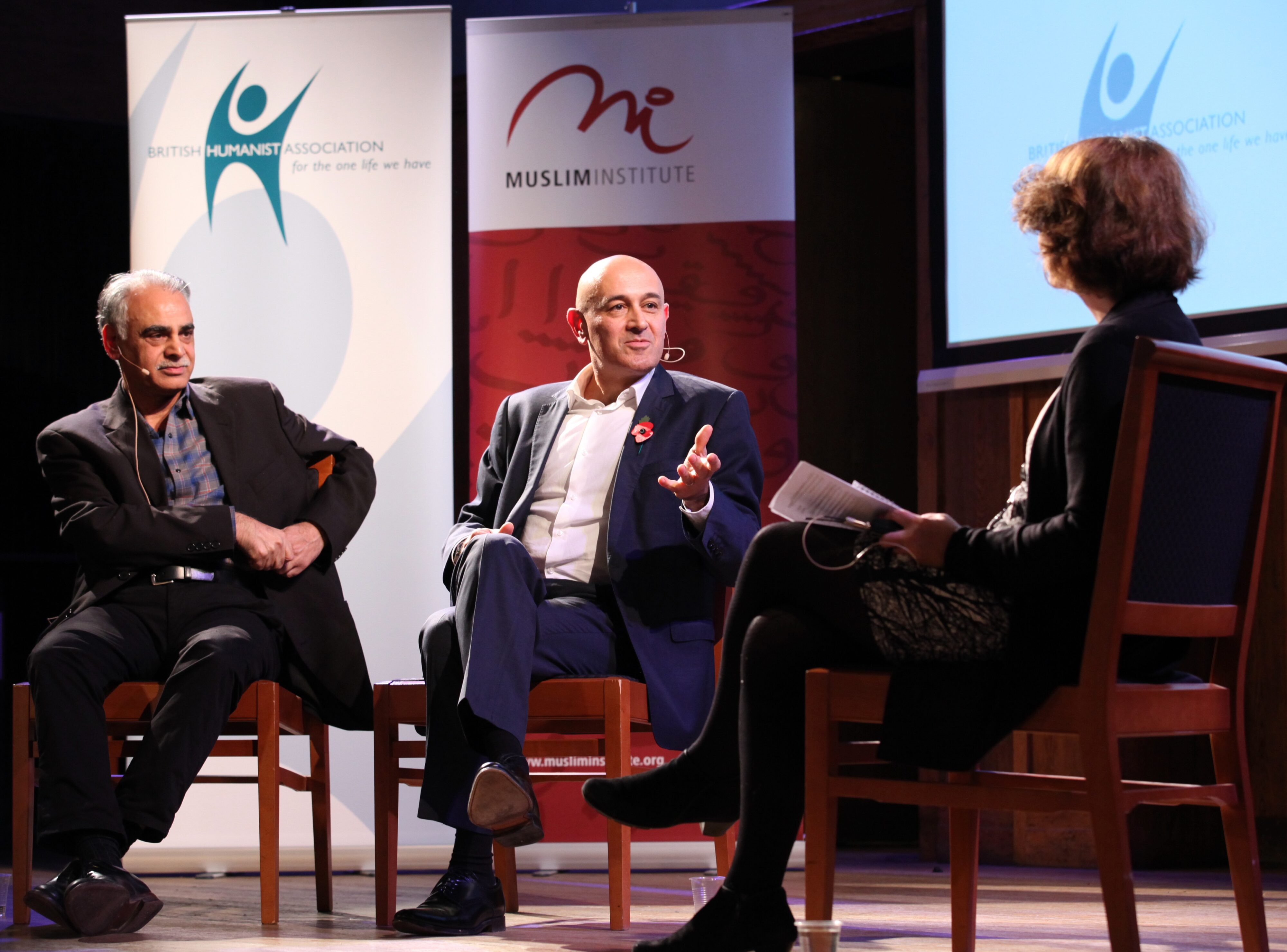 Three people sit on stage having a discussion. Two men sit on the left, one gesturing as he speaks, while a woman on the right listens with papers in hand. Banners for the British Humanist Association and Muslim Institute are behind them.