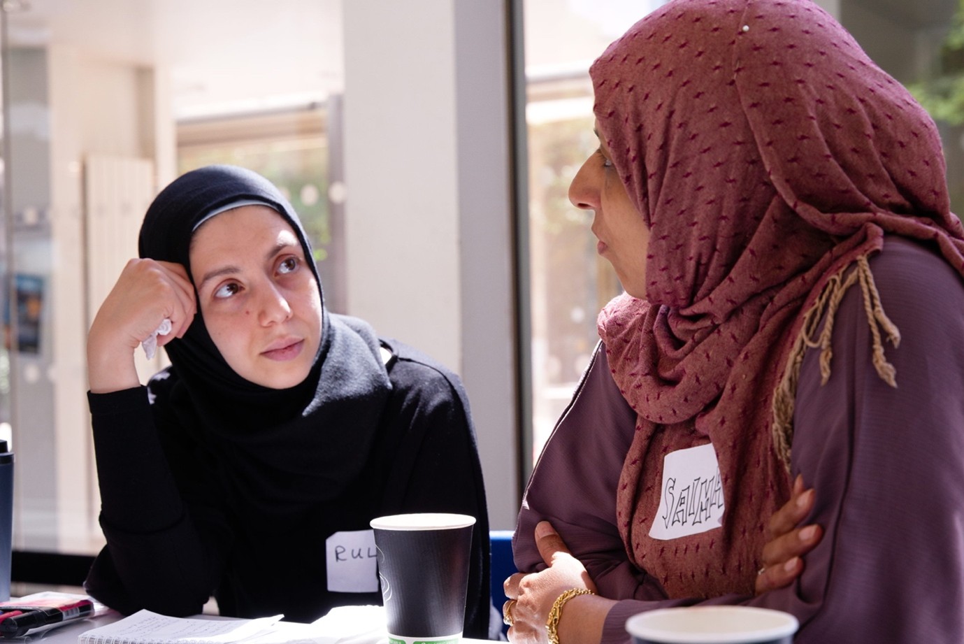 Two women wearing hijabs sit at a table, engaged in conversation. One woman listens attentively with her hand to her face, while the other speaks. Both have name tags; notebooks and cups are on the table.