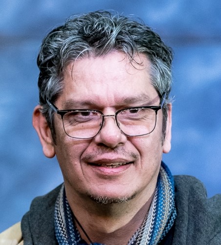 A middle-aged man with gray hair and glasses smiles gently. He is wearing a striped scarf and a jacket, with a blurred blue background behind him.
