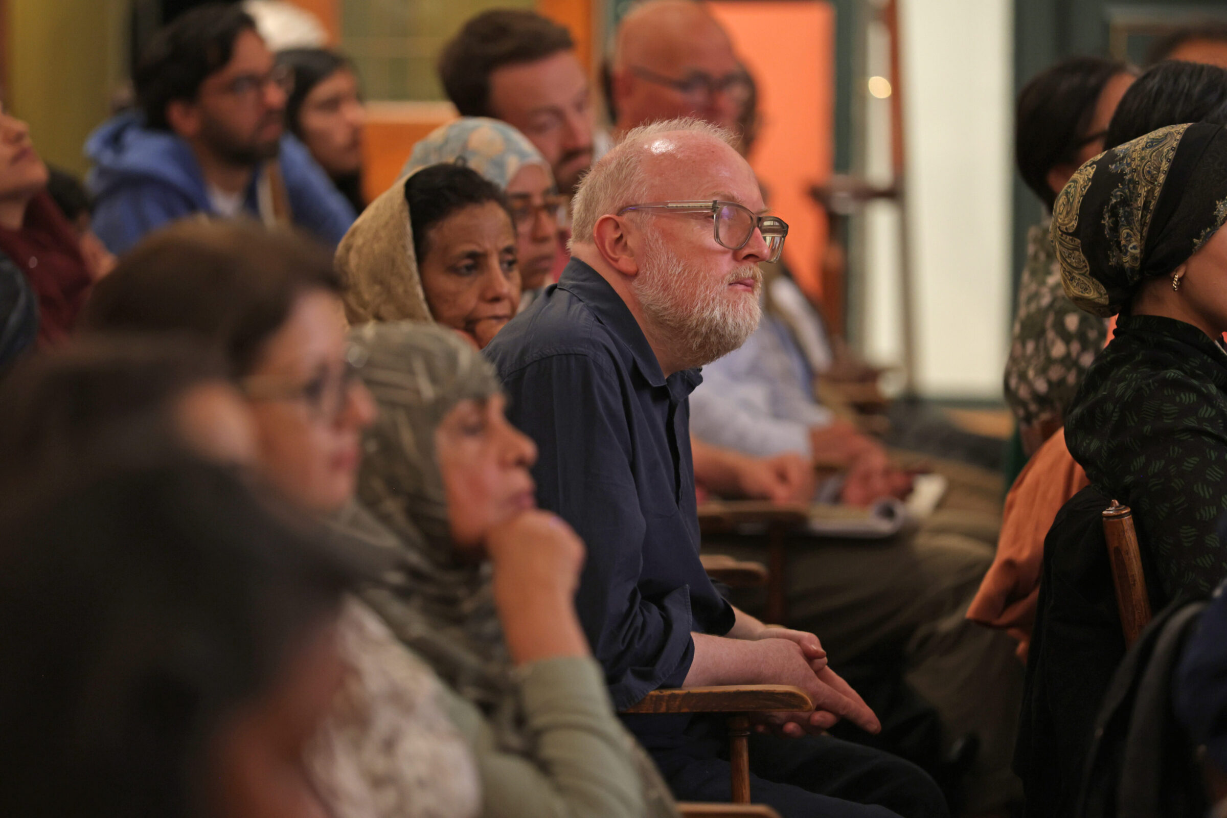 A diverse group of people sit closely together in an audience, listening attentively. A man with glasses and a white beard is in focus among others, all appearing engaged in the event.