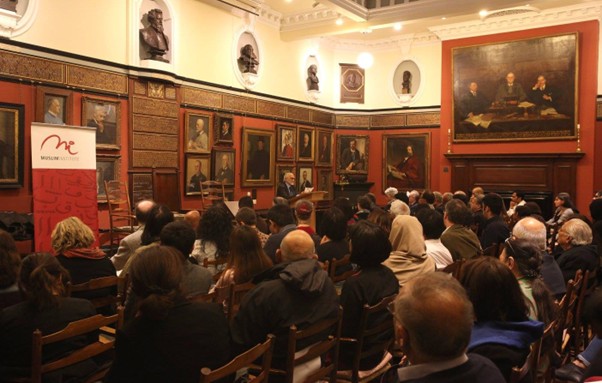 An audience sits in a historic room with ornate decor and large framed portraits on the walls, listening to a speaker at a podium near a red Museum of the Object banner.